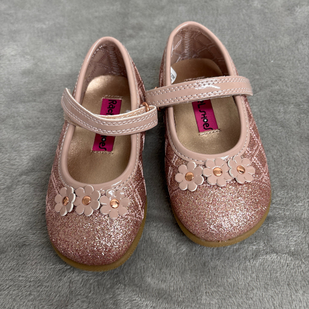 A pair of toddler girl's glitter Mary Jane shoes in a dusty rose pink color, featuring floral appliques and a strap closure, resting on a gray textured surface.