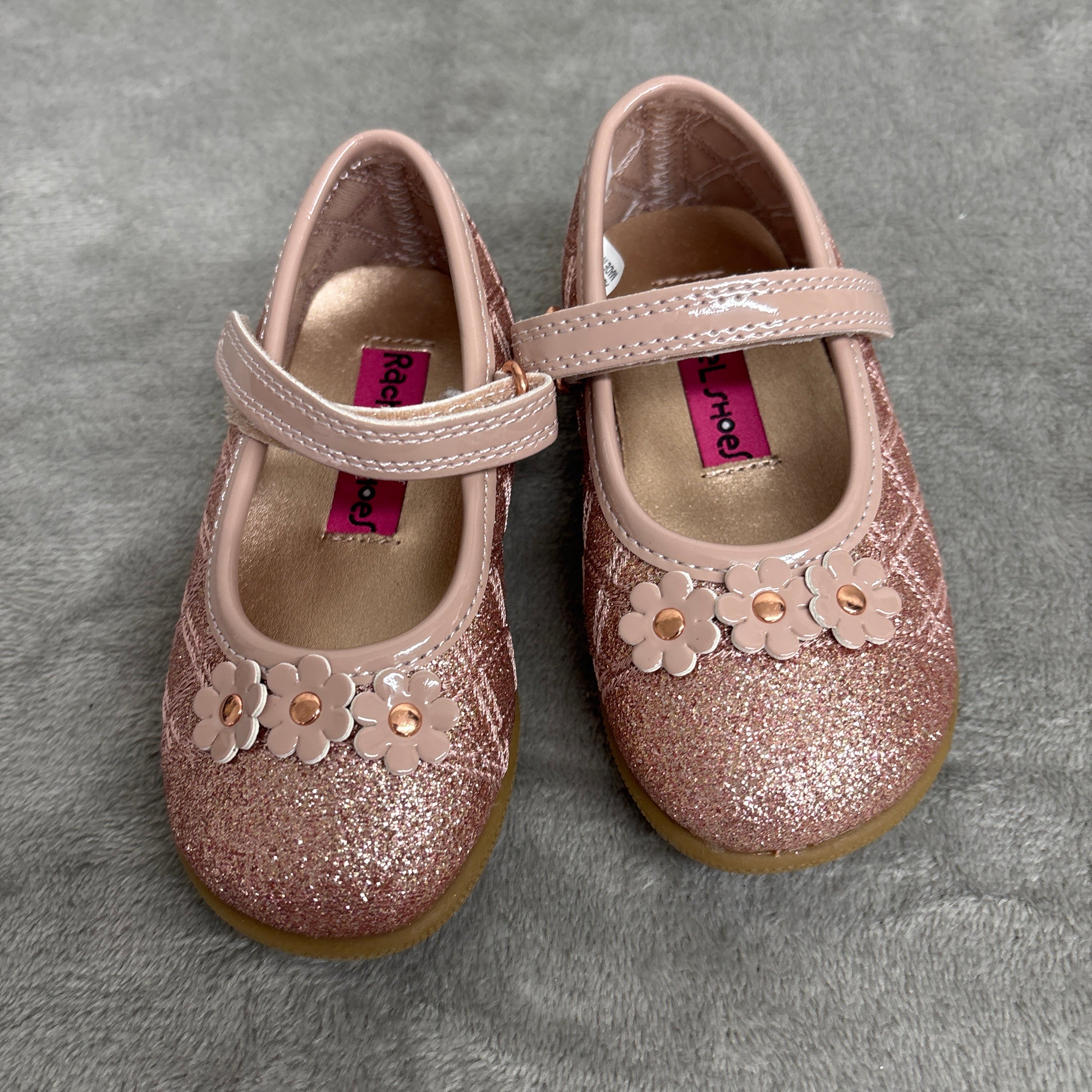 A pair of toddler girl's glitter Mary Jane shoes in a dusty rose pink color, featuring floral appliques and a strap closure, resting on a gray textured surface.
